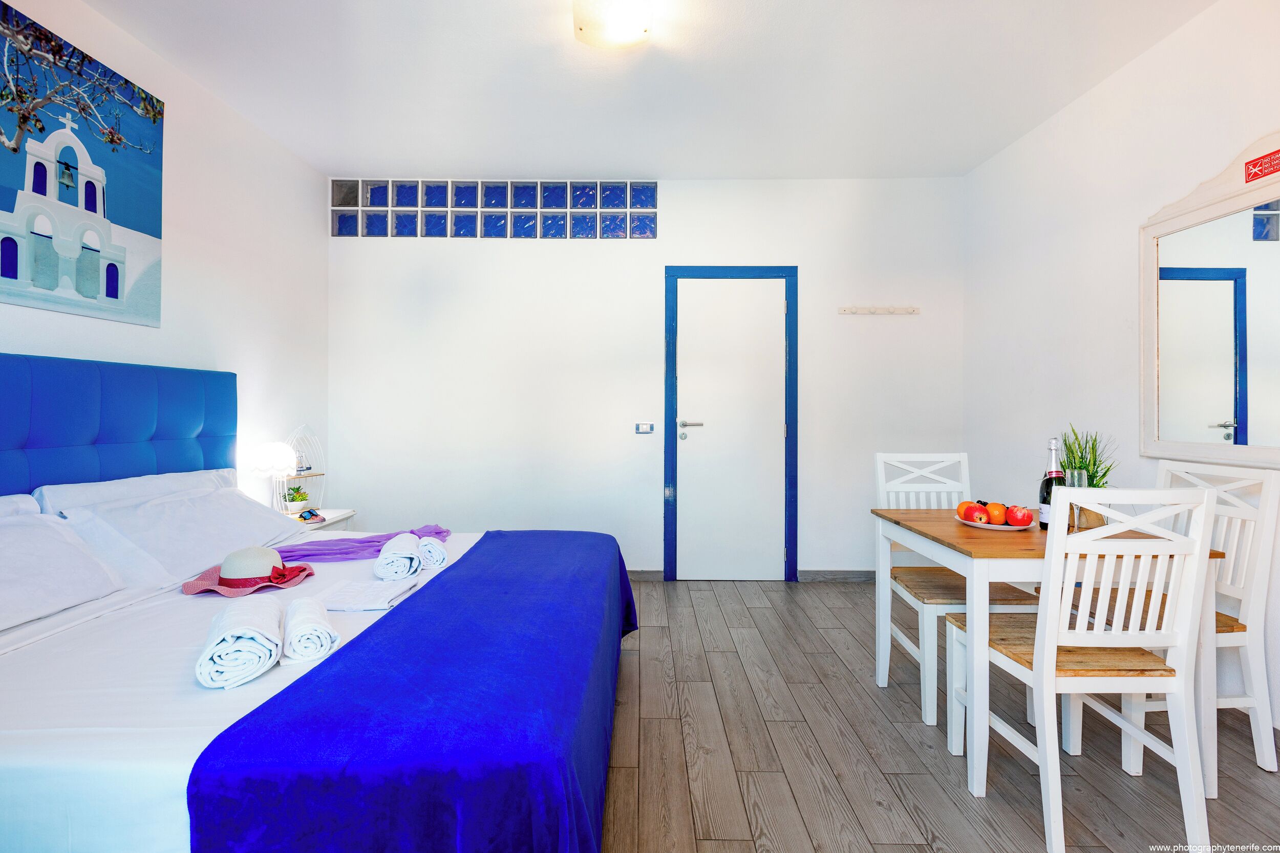 A hotel room with a bed, a desk with a chair, a small table with fruit, and a wall with a blue and white mural.