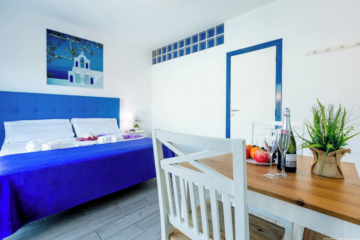 Een hotelkamer met een blauwe spreidek, een wit hoofdeinde, een houten eettafel met een plant erop, en een schilderij van een gebouw aan de muur.