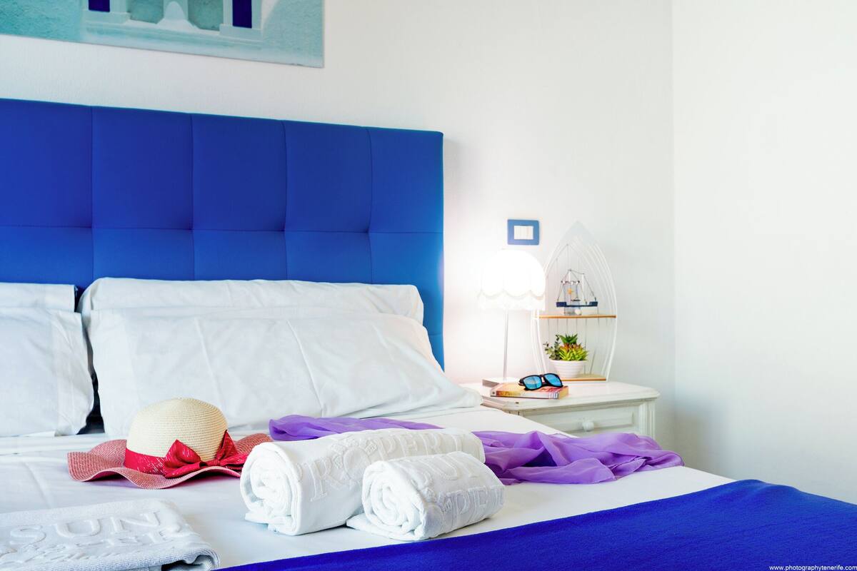 Een net opgemaakt bed met witte lakens, een blauw hoofdbord en een nachtkastje met een lamp, een plant en een paar zonnebrillen.