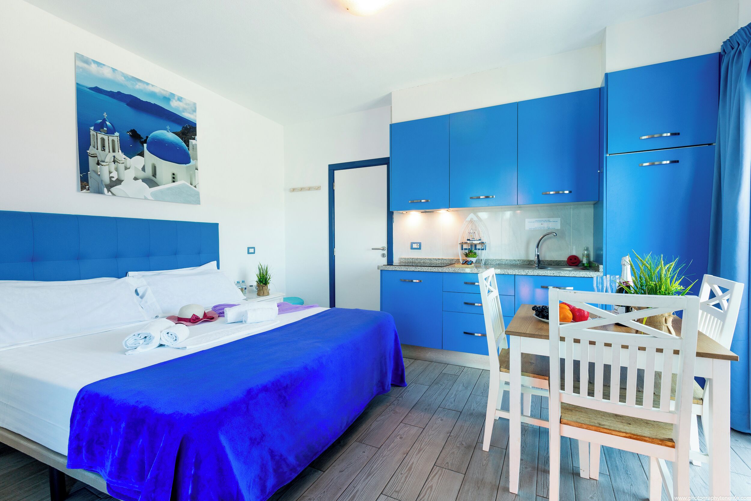 A room with a bed, a dining table with chairs, a kitchen area, and a painting on the wall.