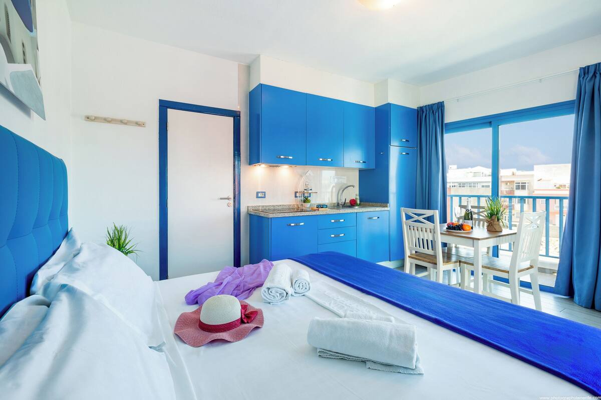 Een moderne hotelkamer met een bed, een klein kitchenette en een balkon met een tafel en stoelen.