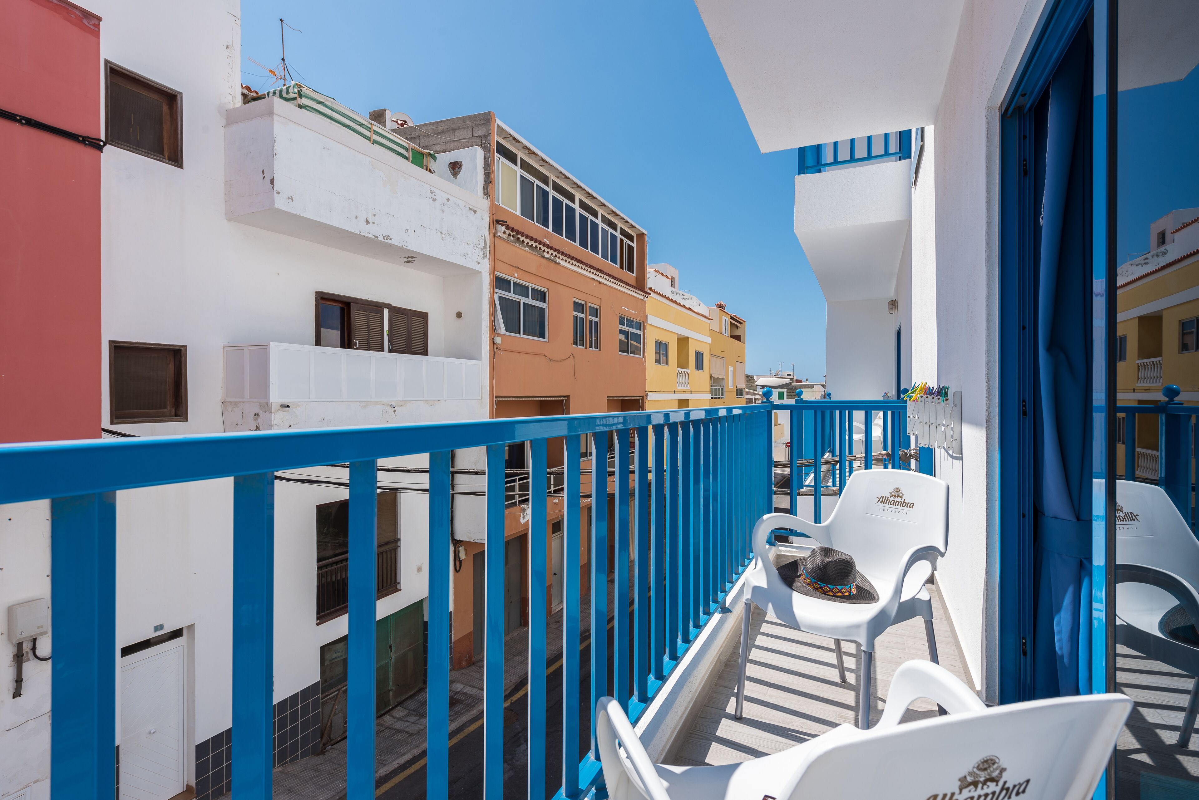 A balcony with white and blue chairs, a white door, and buildings with balconies and windows in the background.