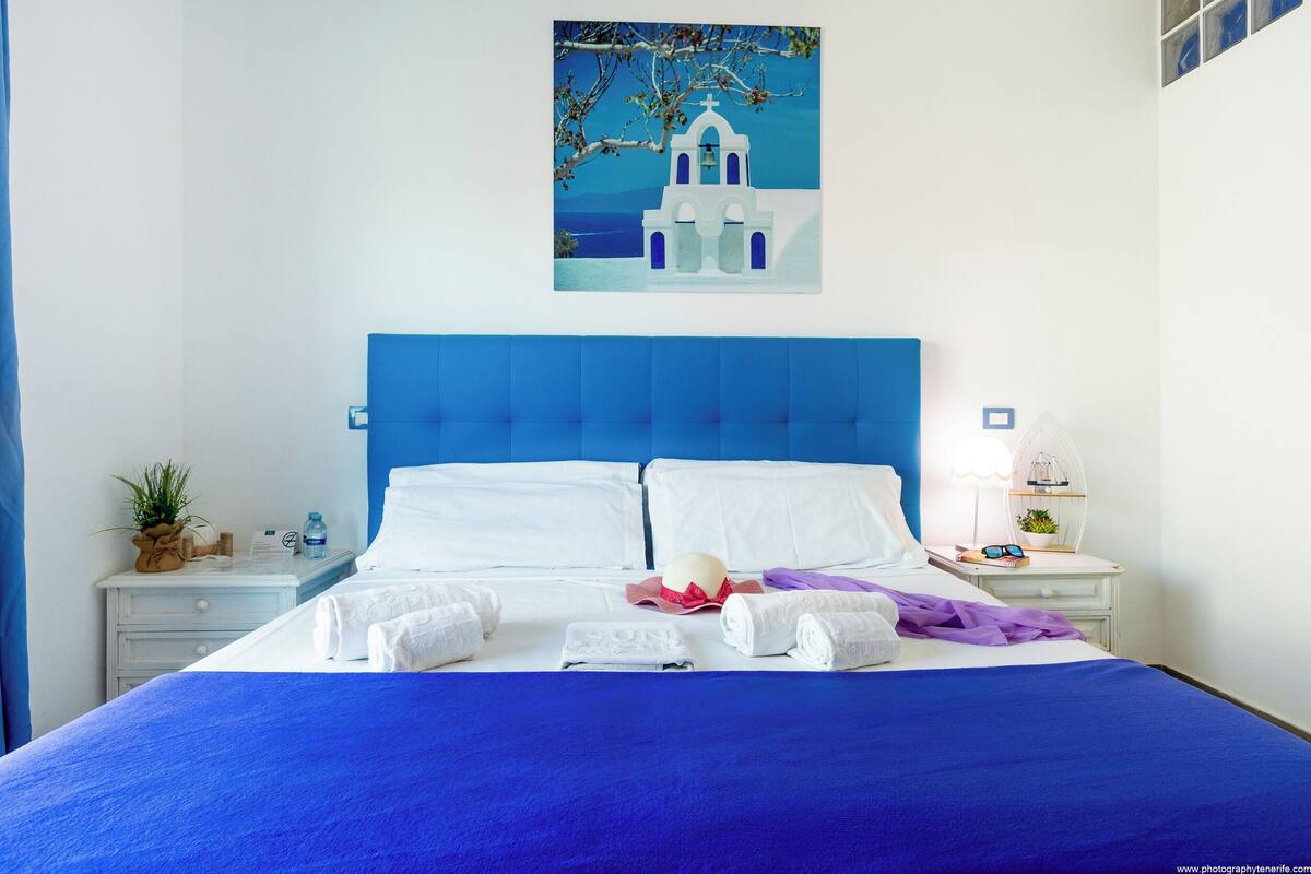 Een netjes opgemaakt bed met witte lakens en een blauw hoofdeinde, een nachtkastje met een lamp en een klein plantje, en een muur met een schilderij van een wit gebouw.