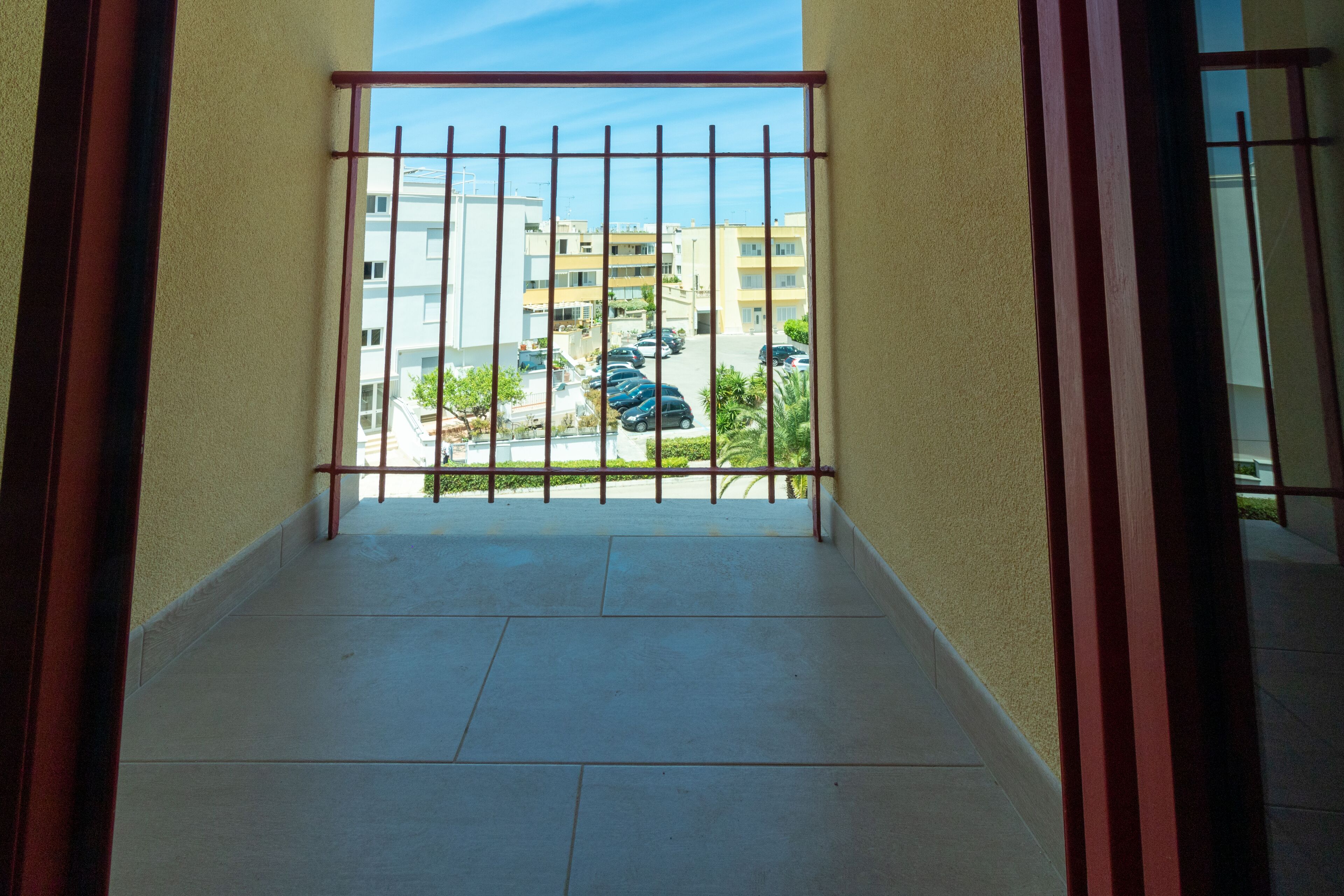 A view from a balcony with a metal gate, leading to a parking area with several cars and buildings in the background.