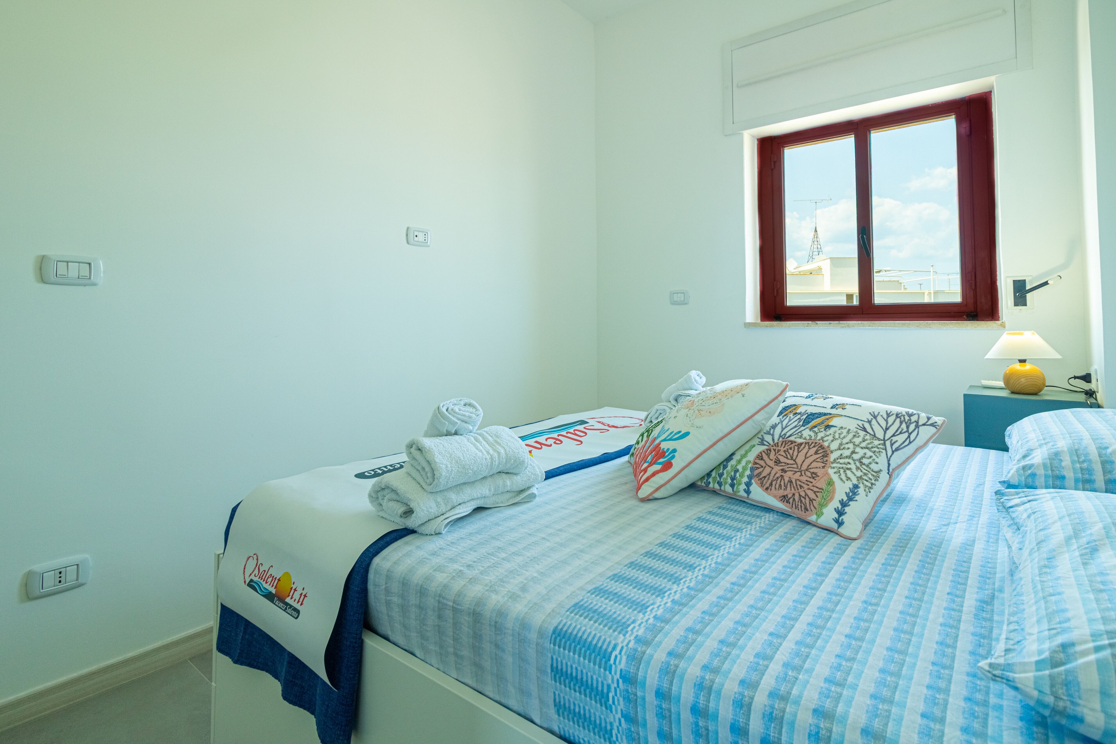 Een netjes opgemaakt bed met een blauw-wit gestreepte dekbedovertrek, een sierkussen met bloemendessin en een opgerolde handdoek aan de zijkant.