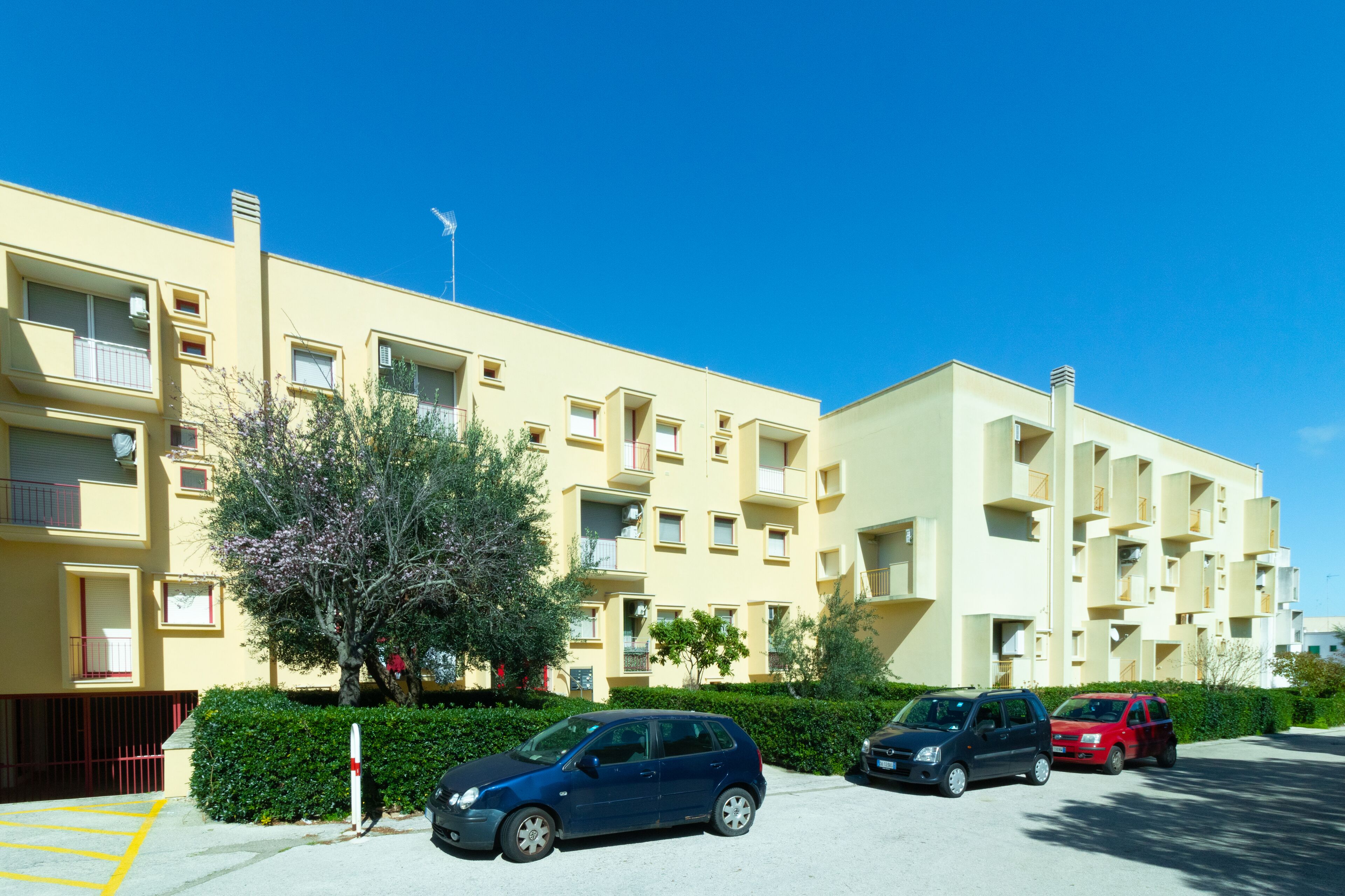 Un edificio residencial de varios pisos con balcones, un estacionamiento y un cielo azul despejado.