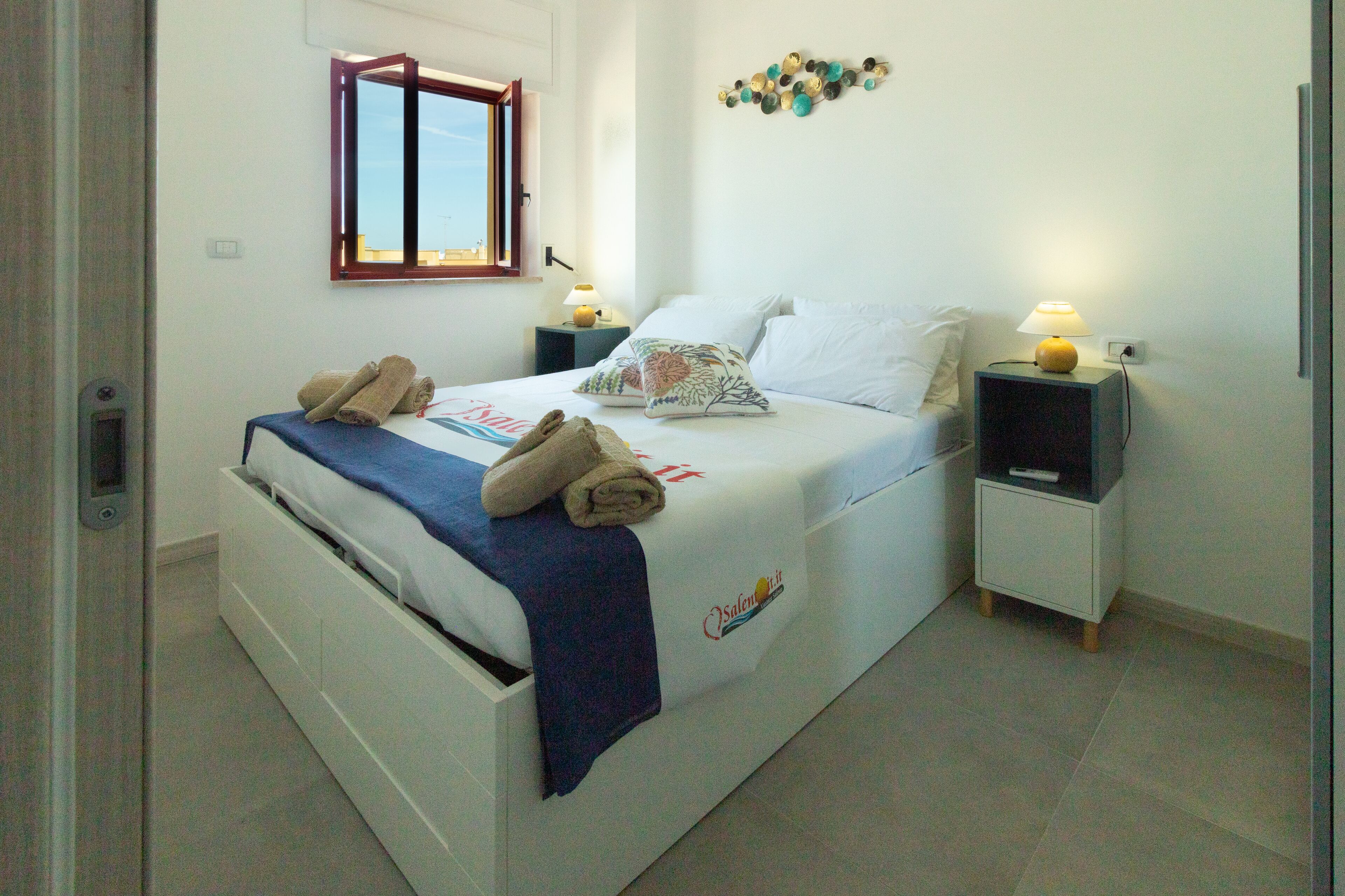 Cama bem arrumada com toalhas, mesa de cabeceira com abajur e um detalhe decorativo na parede.