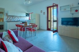 A modern living room with a purple sofa, a dining area with red chairs, a kitchen with white cabinets, and a TV on a wooden stand.