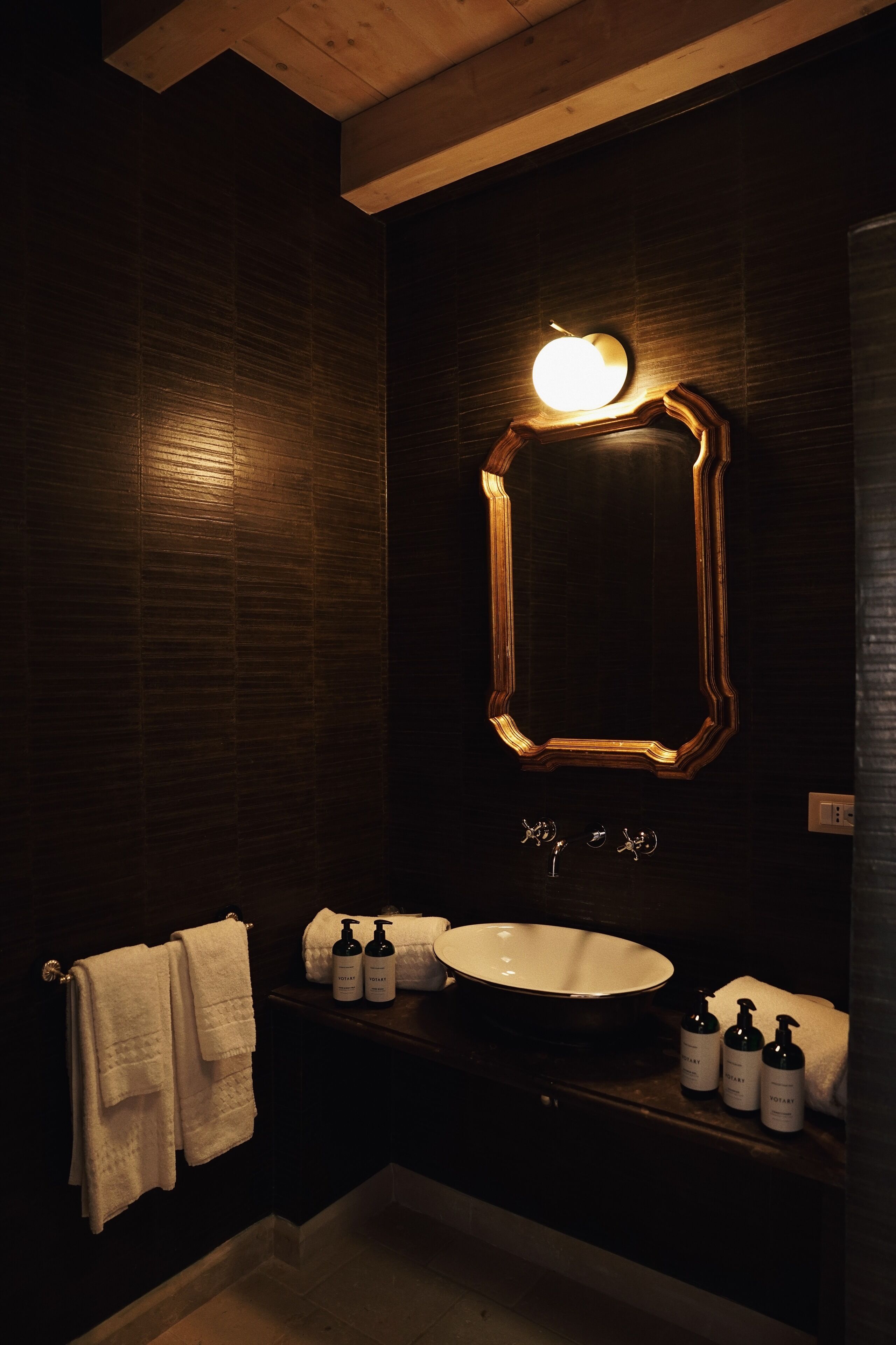 Un baño de estilo oscuro con un espejo grande, un lavabo blanco y dos botellas de champú.