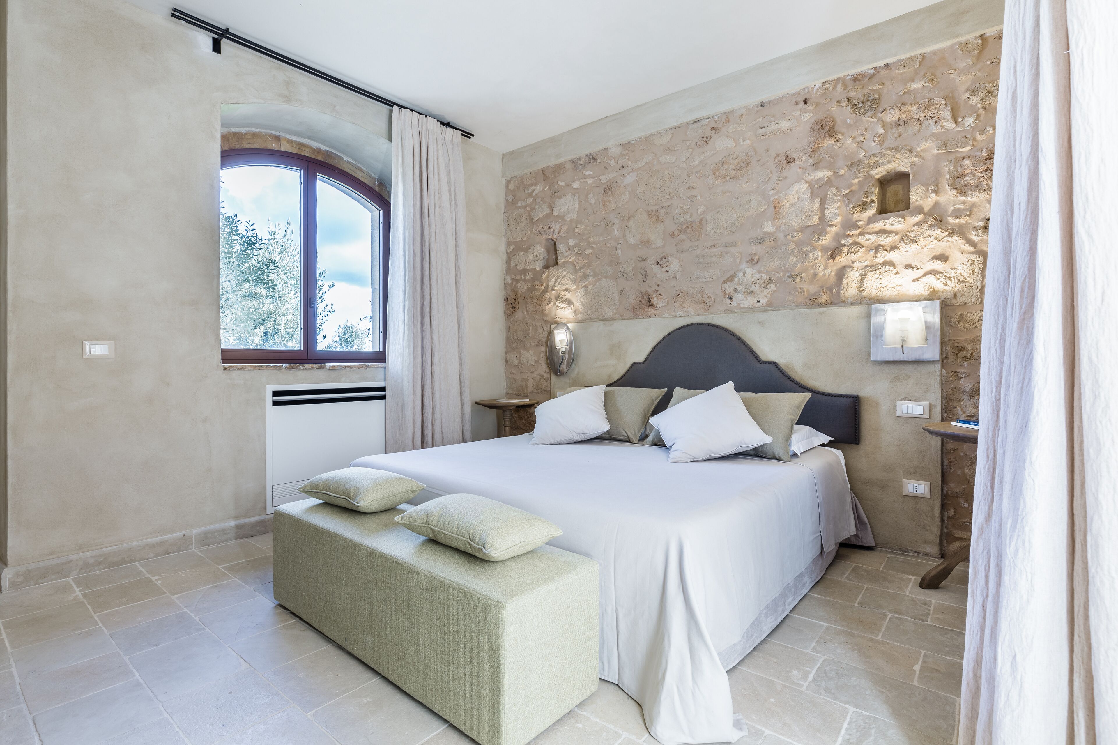 A bedroom with a stone wall, a large bed with white bedding, a bench, and a window with curtains.