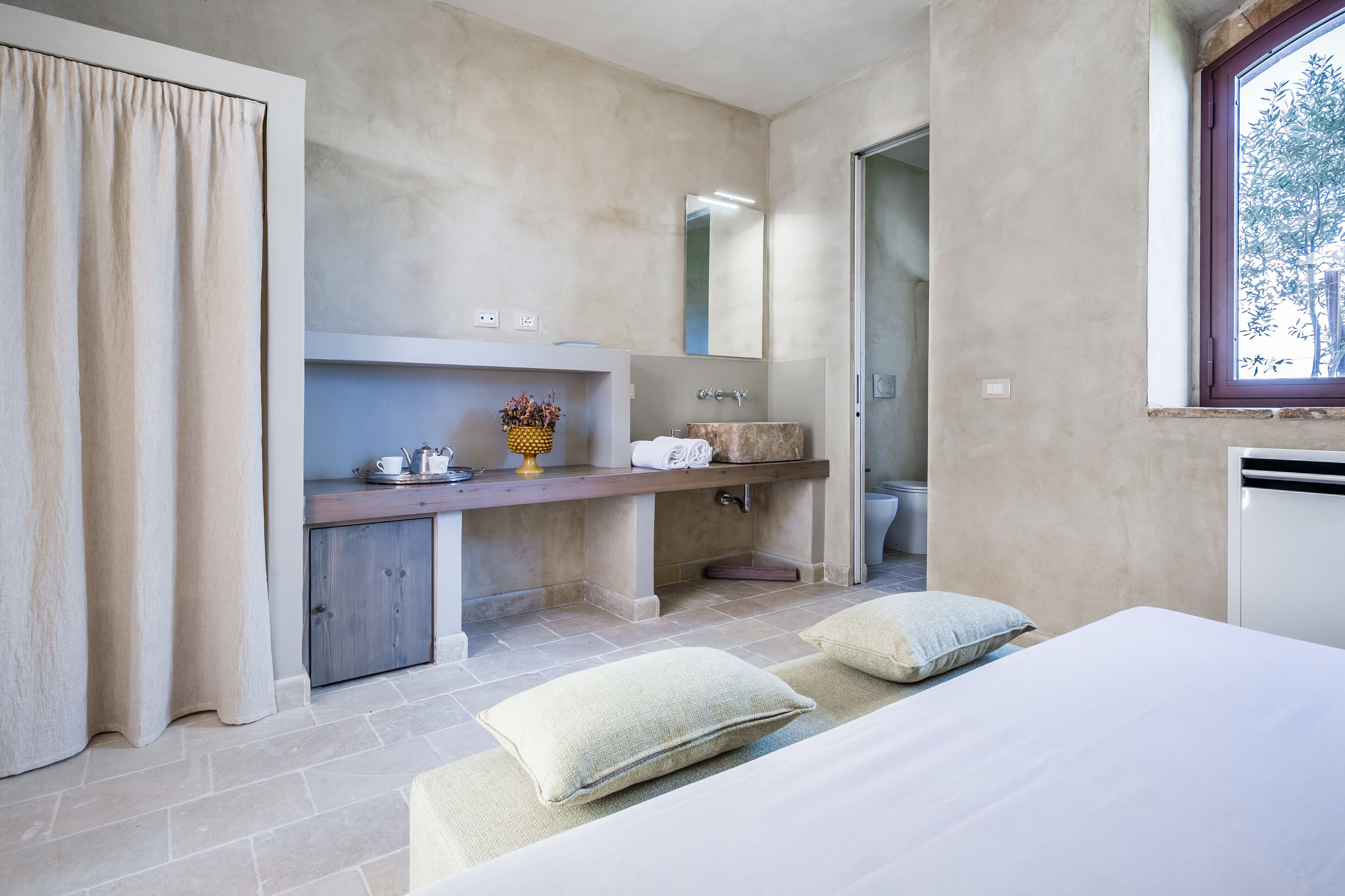 A modern bathroom with a sink, mirror, and toilet. There is a bed with pillows and a window with a view of trees.