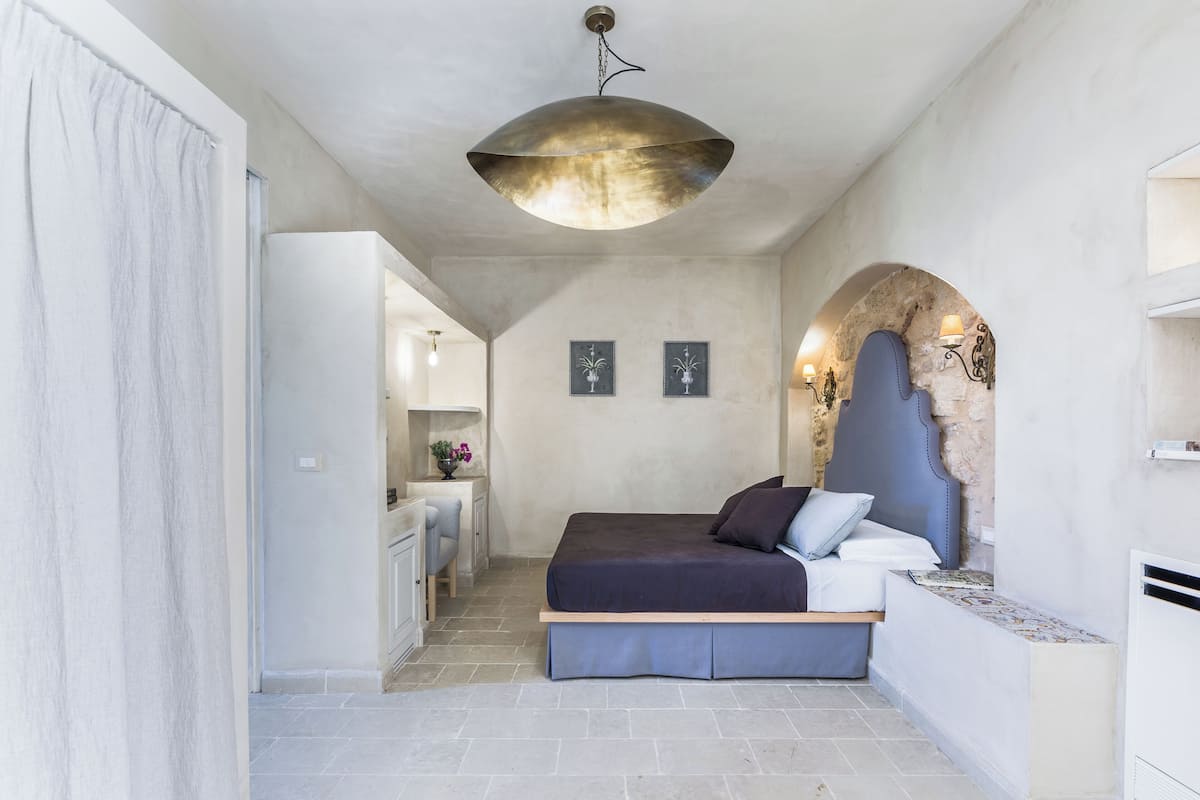 Quarto com cama, luminária pendente, arco de pedra e pequena escrivaninha com cadeira.