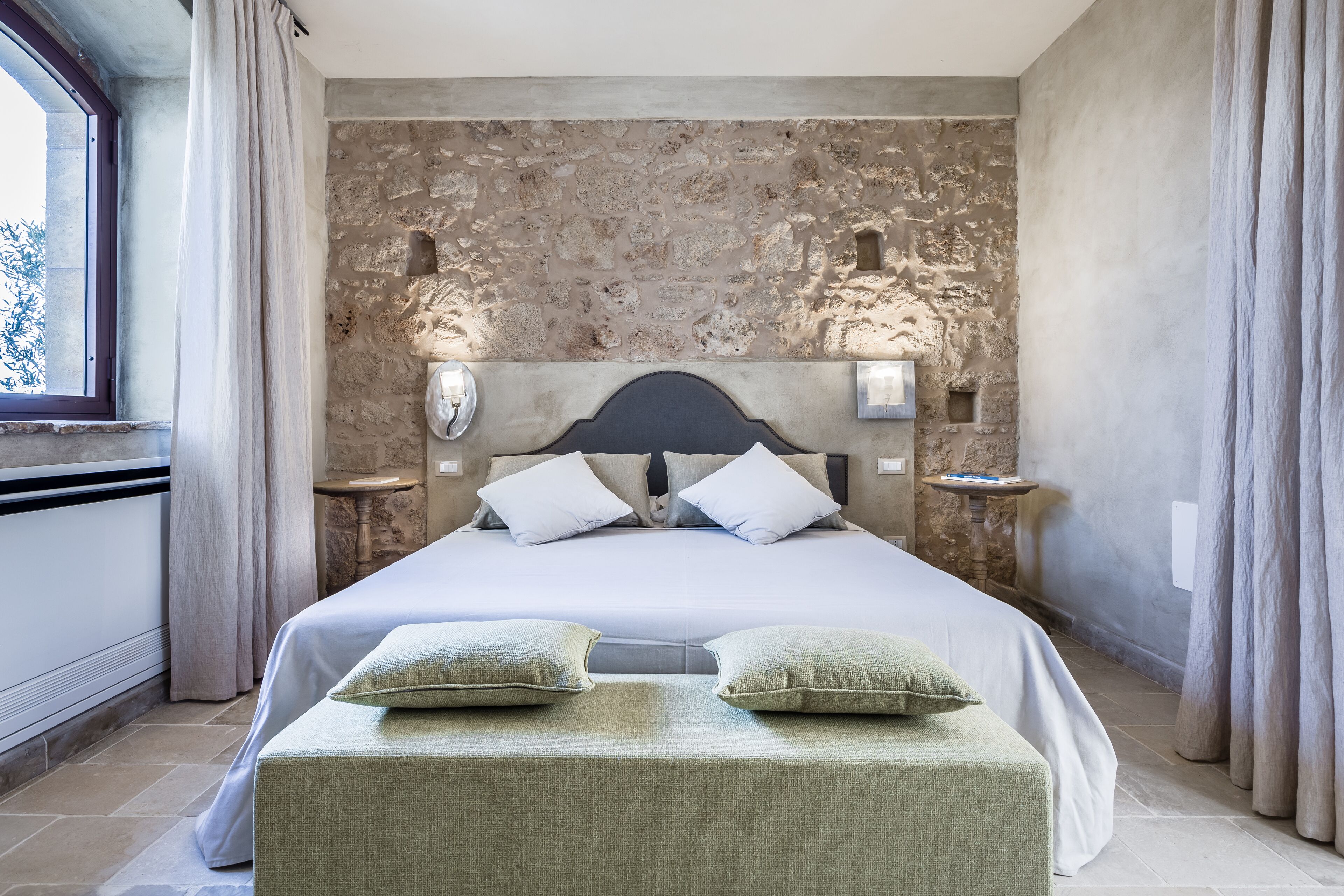 Una cama grande con un cabecero de pared de piedra, dos mesitas de noche con lámparas y vistas al exterior a través de una ventana.