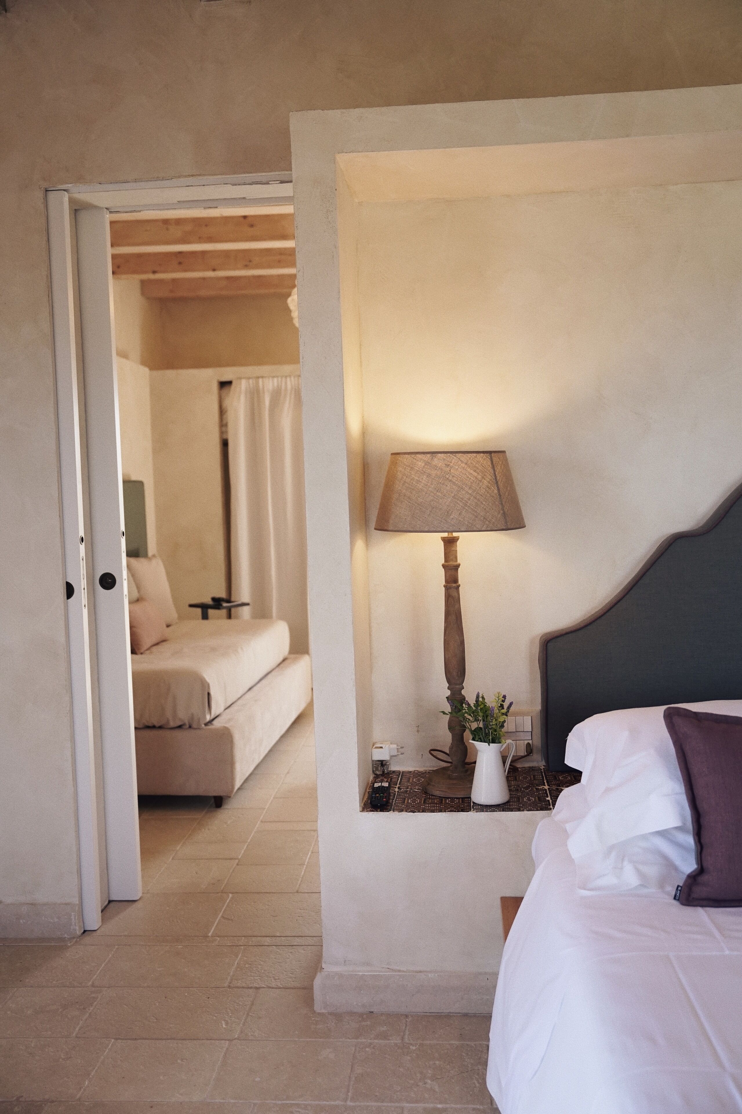 A hotel room with a bed, a nightstand with a lamp, a small table with a vase, and a view into another room.
