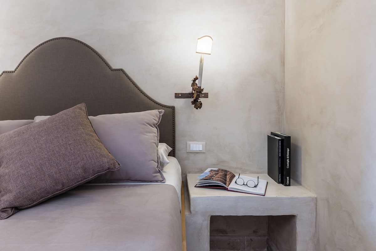 Uma cama bem arrumada com cabeceira, mesa de cabeceira com abajur, um livro e um par de óculos.