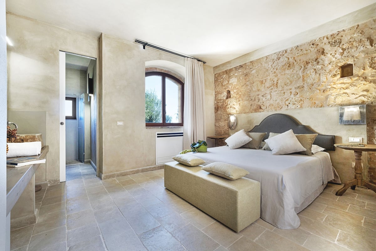 Quarto moderno com uma cama grande, um banco e uma parede de pedra.
