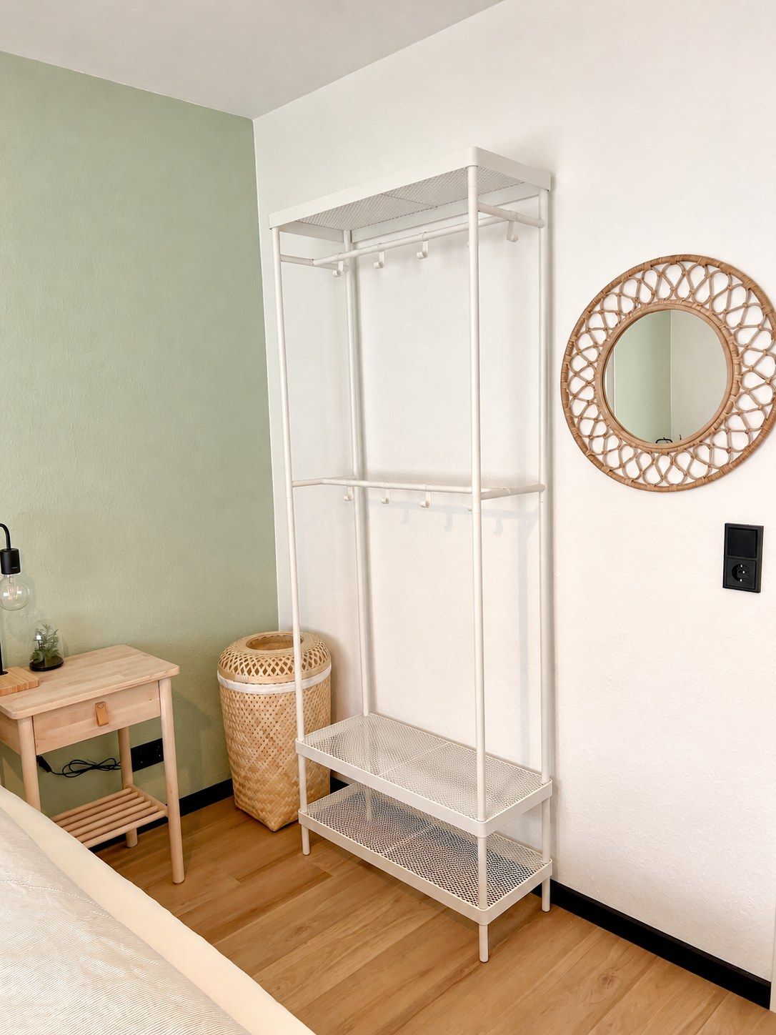 Habitació amb una estant metàl·lica blanca, un mirall rodó, una taula de fusta petita amb una làmpada i una cistella de vimet.