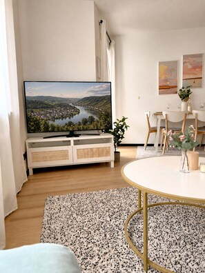 A living room with a flat-screen TV, a dining table with chairs, a white cabinet, a plant, and a rug. - JUPAS° Boho Home – Balcony, Parking & Playground (Trier)