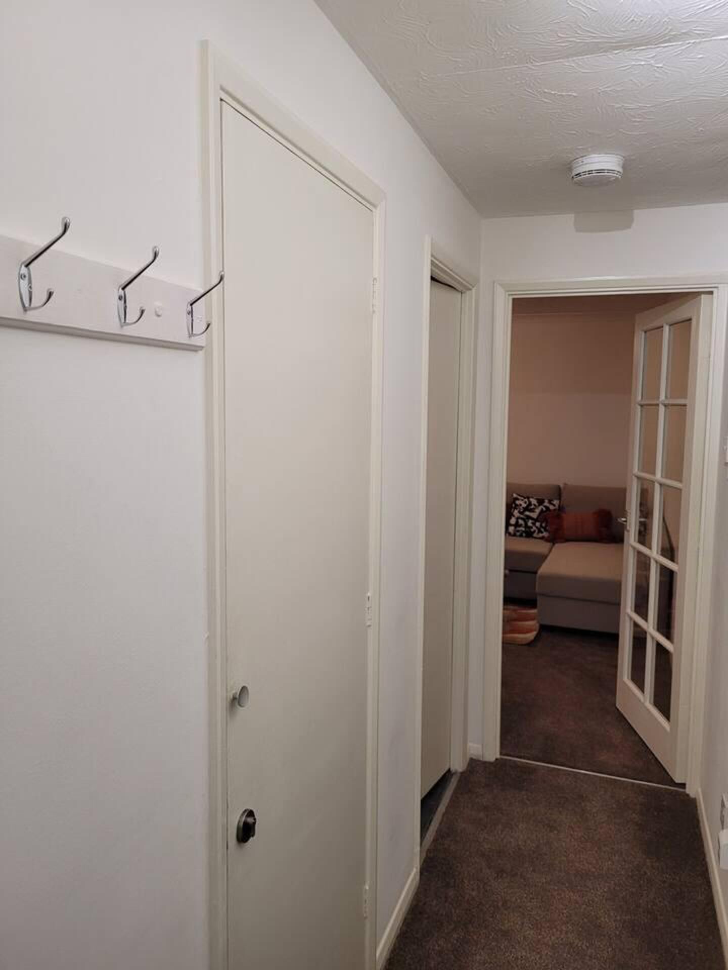 Un couloir avec un porte-manteau, une porte avec un trou de serrure et une pièce avec un canapé et des coussins.