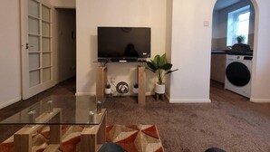A living room with a TV, a glass coffee table, a plant, and a washing machine in the background. - Tastefully Decorated 1 bed Flat Near Abbeywood (Belvedere)