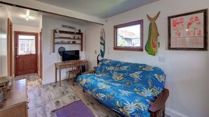 A living room with a wooden desk, a television, a blue sofa with a tropical print, a surfboard, and a framed poster on the wall. - Latitude Adjustment - Ocracoke Island (Ocracoke)