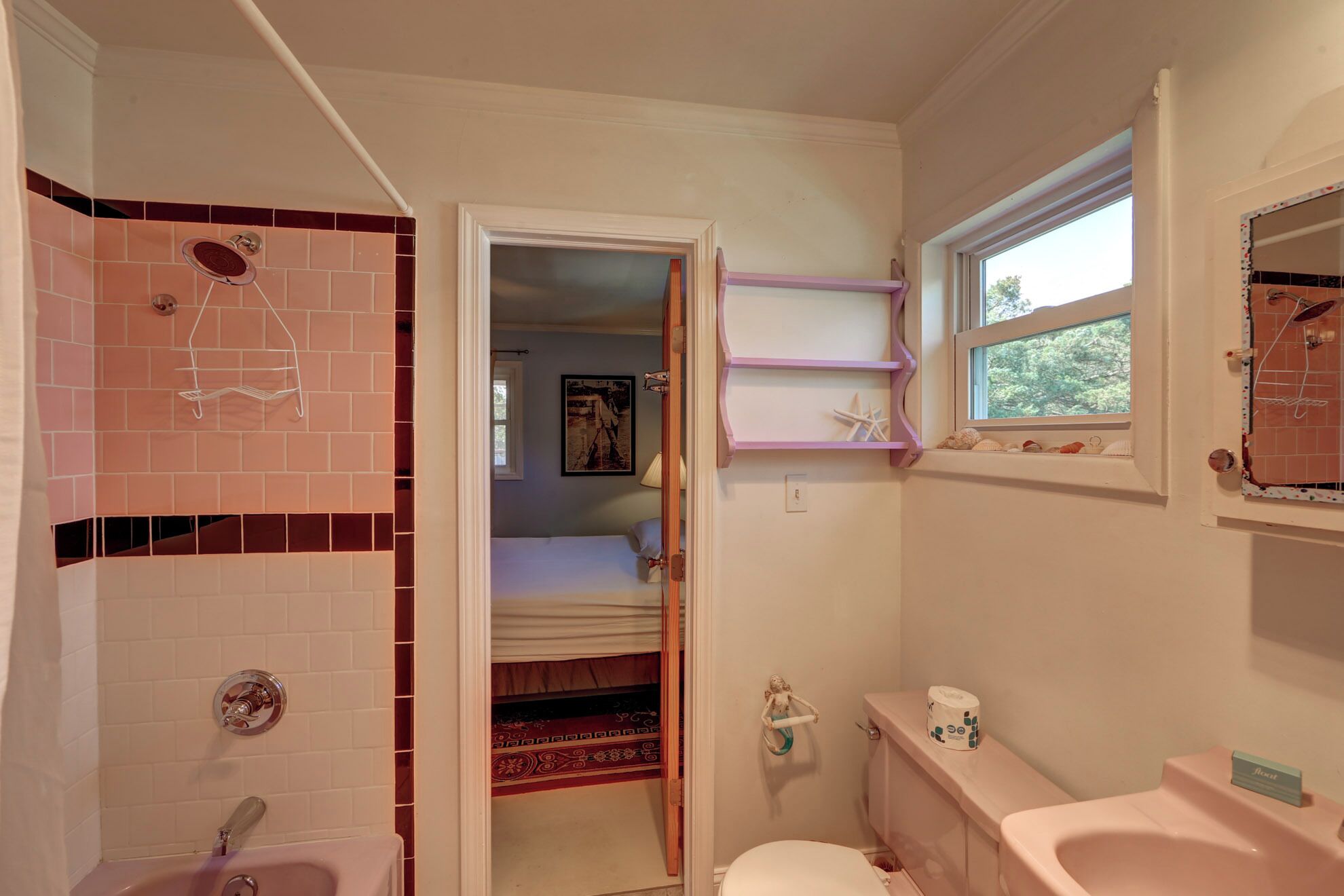 Une salle de bain compacte comprenant une douche, des toilettes et un lavabo. Une fenêtre laisse entrer la lumière naturelle et une petite étagère est décorée de quelques objets.