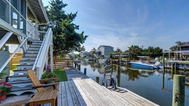 Une terrasse en bois avec un banc et une chaise, donnant sur une étendue d’eau calme, avec des bateaux et des maisons en arrière-plan.