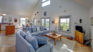 A spacious living room with a high ceiling, a dining area, and a kitchen. There are large windows providing natural light and views of the outdoors. - Heart's Desire - Ocracoke Island (Ocracoke)