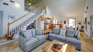 A spacious living area with a staircase leading to a kitchen, featuring a grey striped sofa, a wooden coffee table, and a dining area with chairs. - Heart's Desire - Ocracoke Island (Ocracoke)