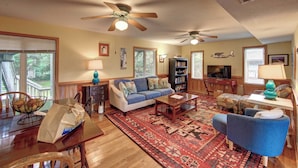 A spacious living room with a blue sofa, a wooden coffee table, and a patterned rug. There are two ceiling fans, a bookshelf, and a television. - Siren Song - Ocracoke Island (Ocracoke)