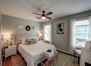 Une chambre à coucher comprenant un lit, un ventilateur de plafond, une fenêtre avec des stores, une chaise et une table de chevet avec une lampe.