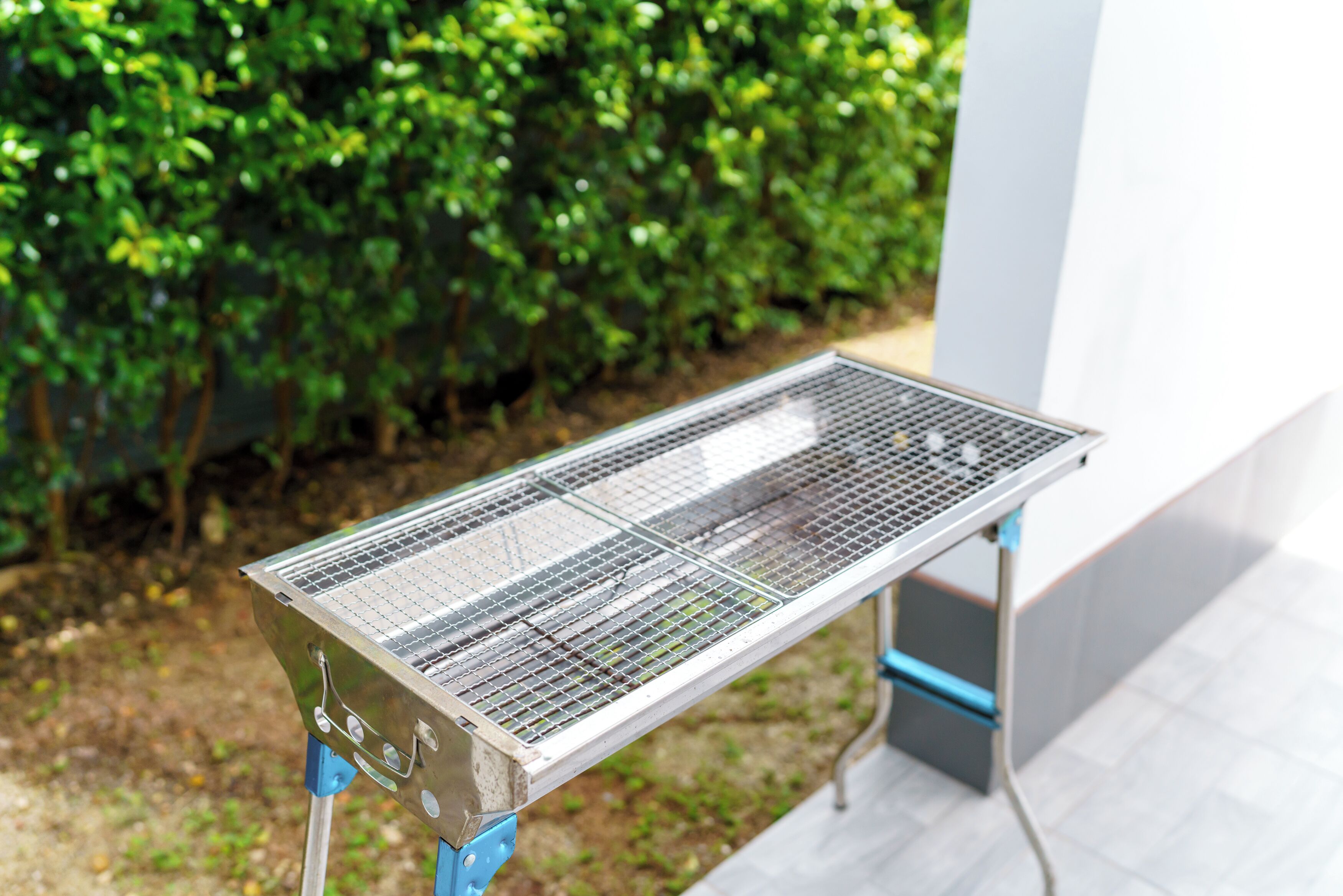 A portable grill with a metal grate, set up outdoors with a green hedge in the background.