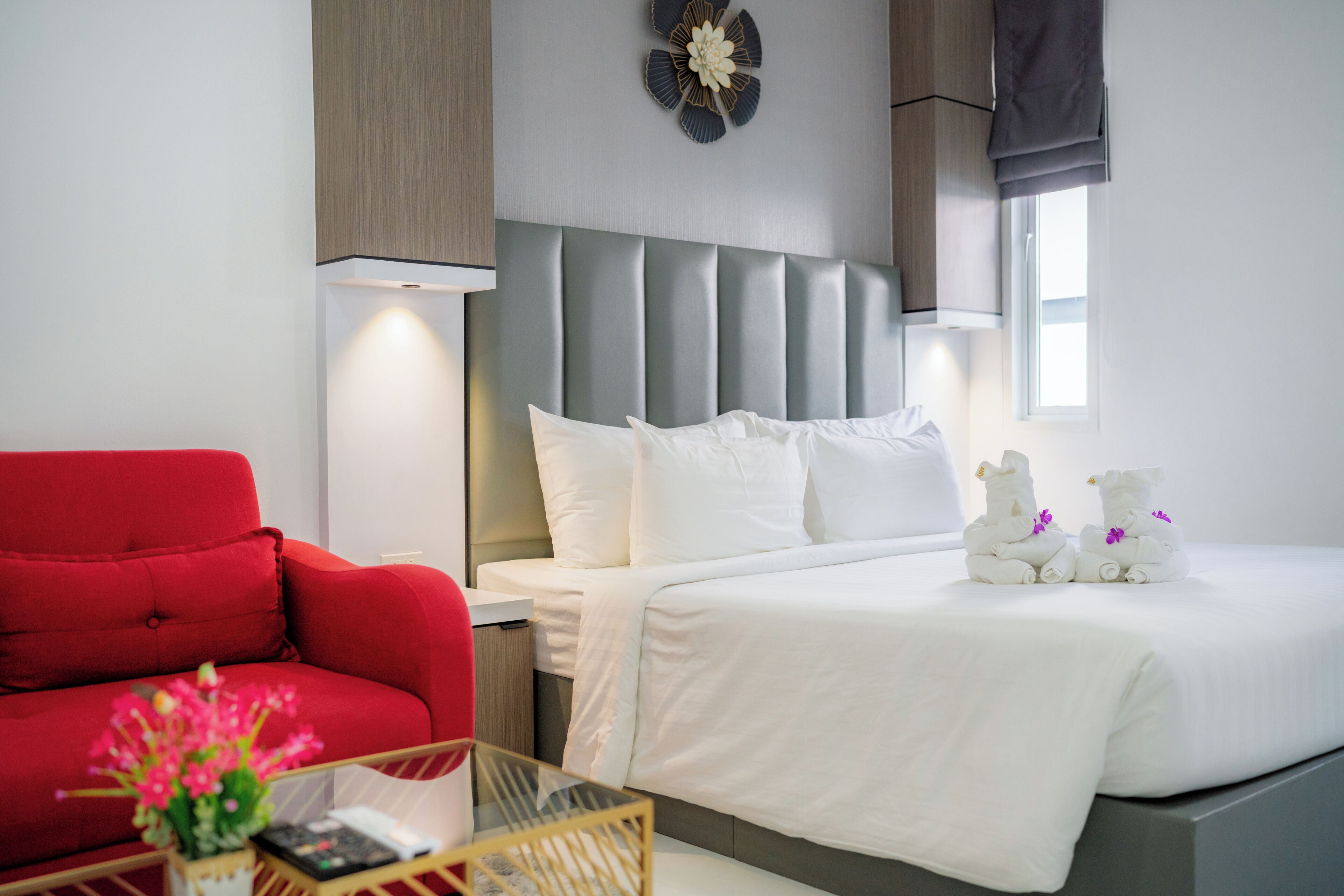 A hotel room with a bed, a red armchair, a bedside table with a vase of flowers, and a wall clock.