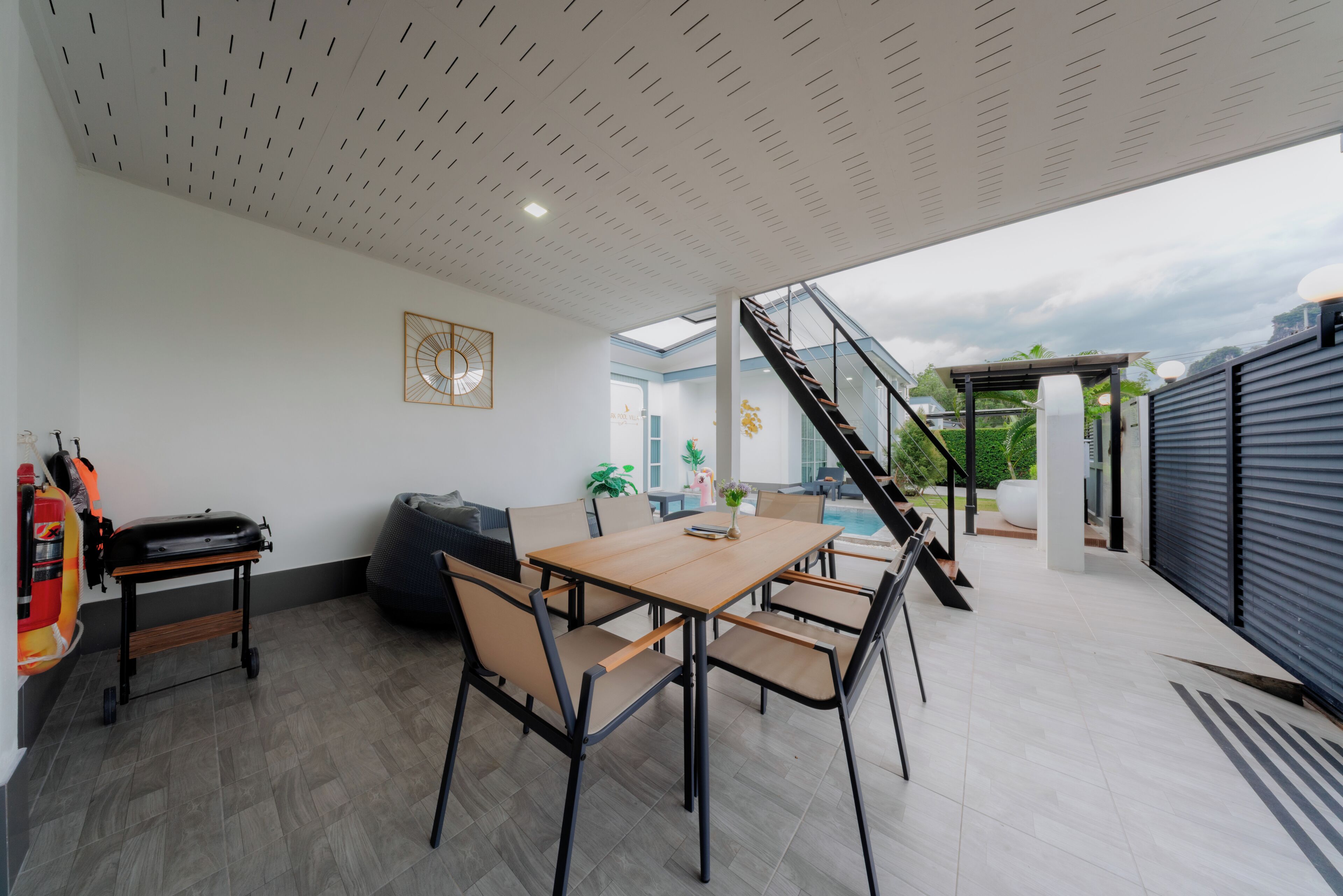A modern outdoor dining area with a wooden table and chairs, a fire extinguisher, and a staircase leading to a pool area.