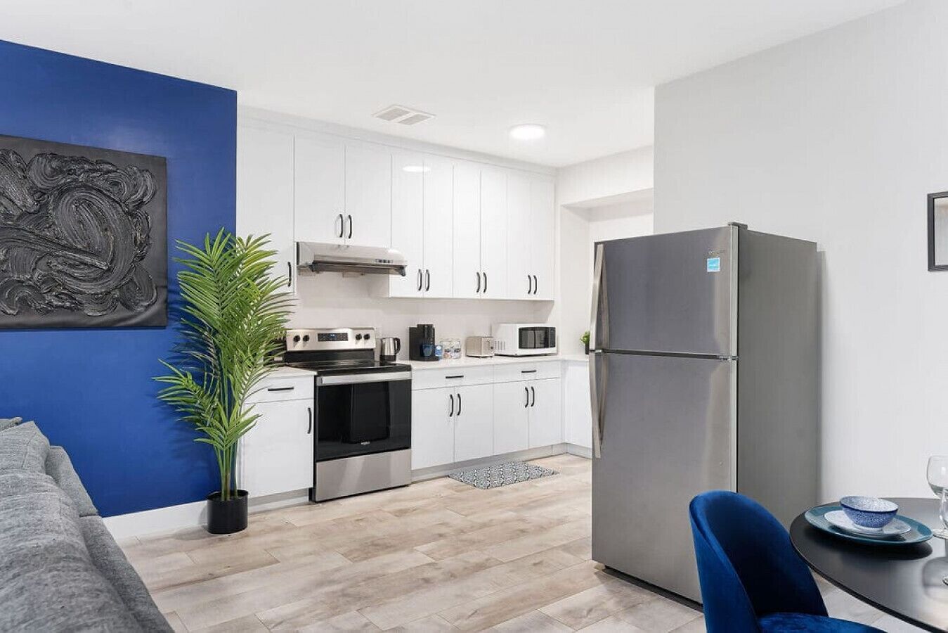 A modern kitchen with white cabinets, stainless steel appliances, a blue accent wall, and a dining area with a blue chair.