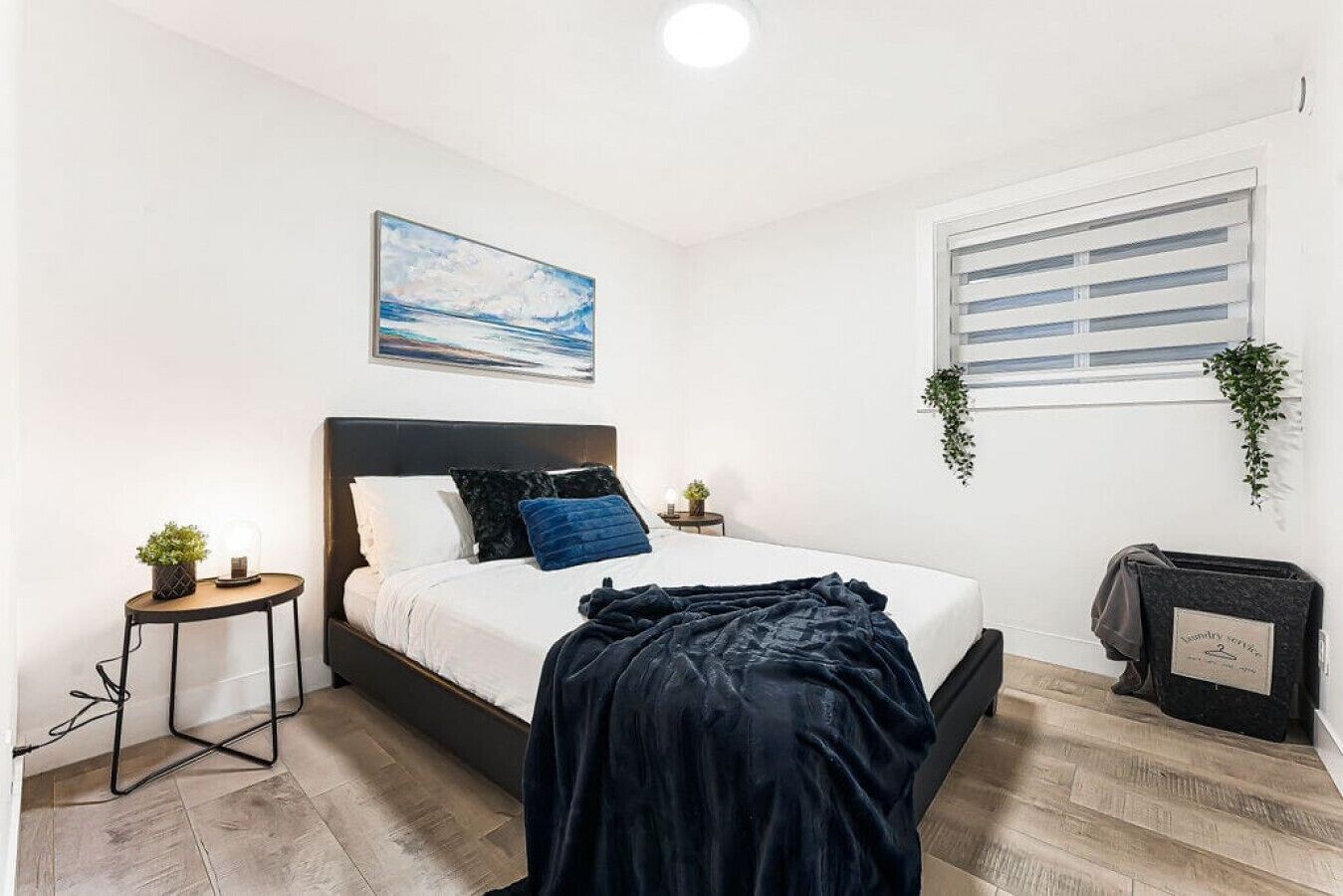 Une chambre à coucher moderne avec un lit, une table de chevet, une petite table avec une plante, un tableau au mur, une fenêtre avec des stores et un panier à linge.