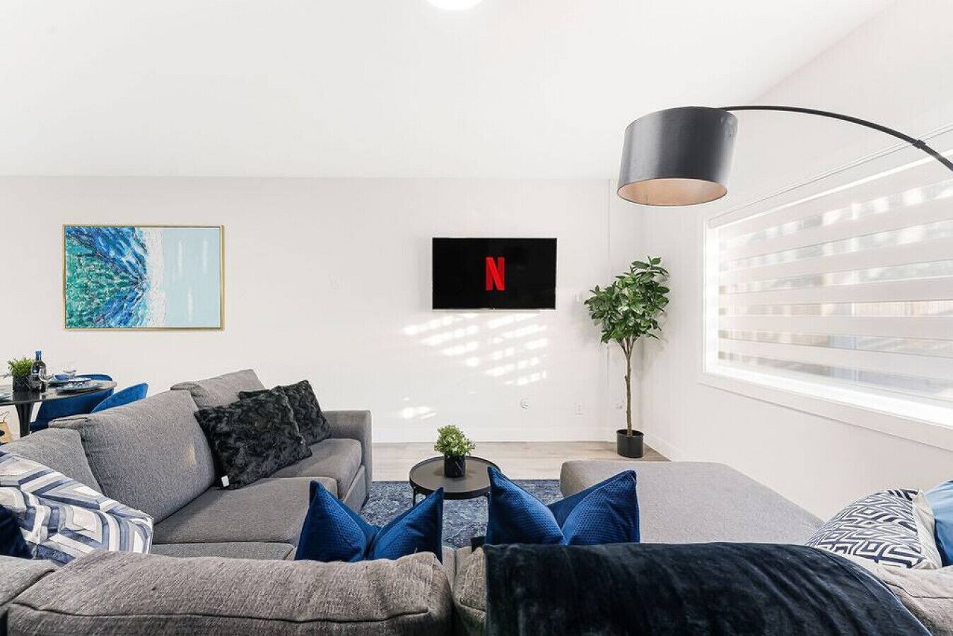 Una sala de estar moderna con un sofá gris, cojines azules, una mesa de centro redonda, una lámpara de pie negra, un cuadro abstracto enmarcado, el logotipo de Netflix, una planta en maceta y una ventana con persianas.