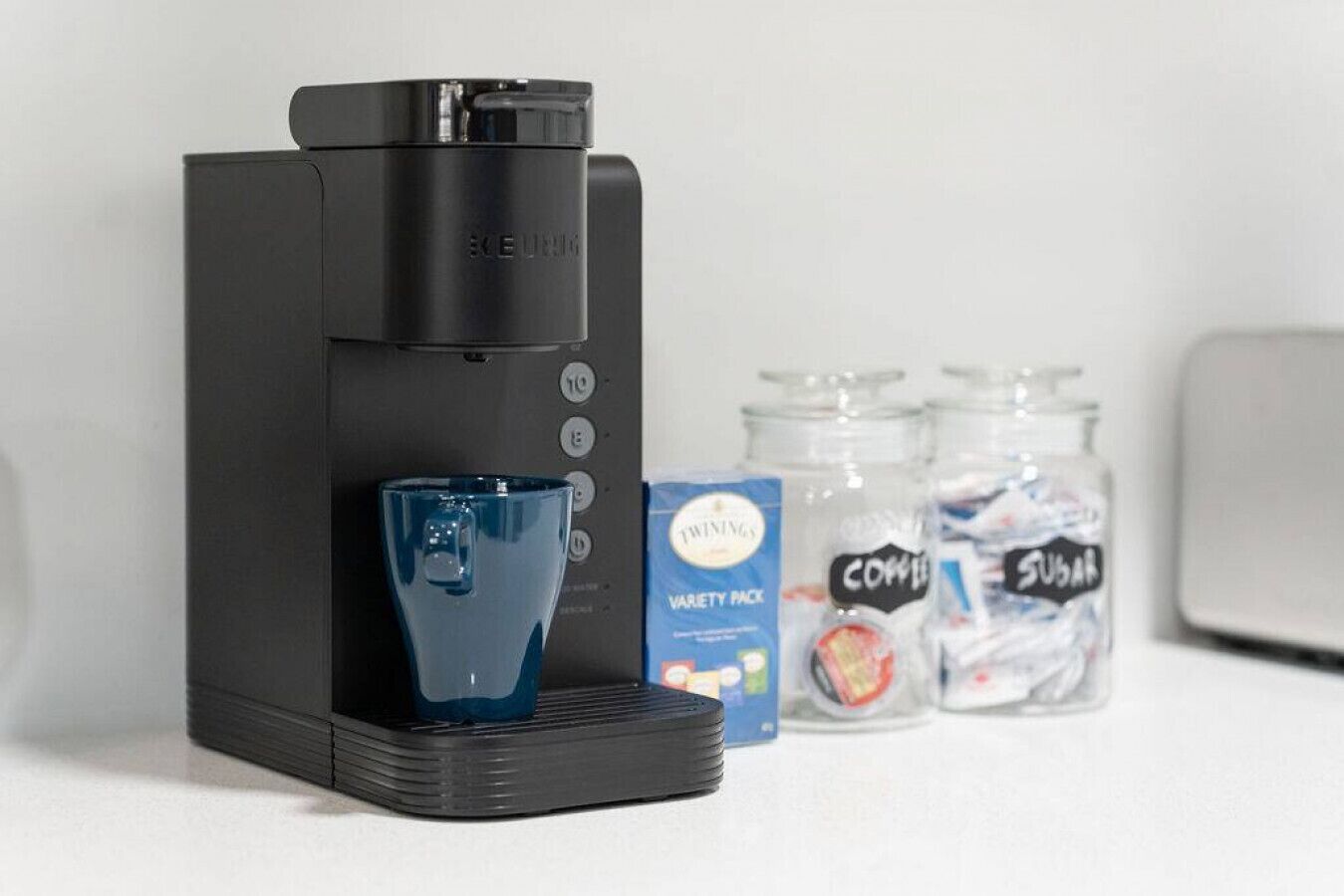 A Keurig coffee maker with a blue cup, a variety pack, and two jars labeled 'coffee' and 'sugar'.