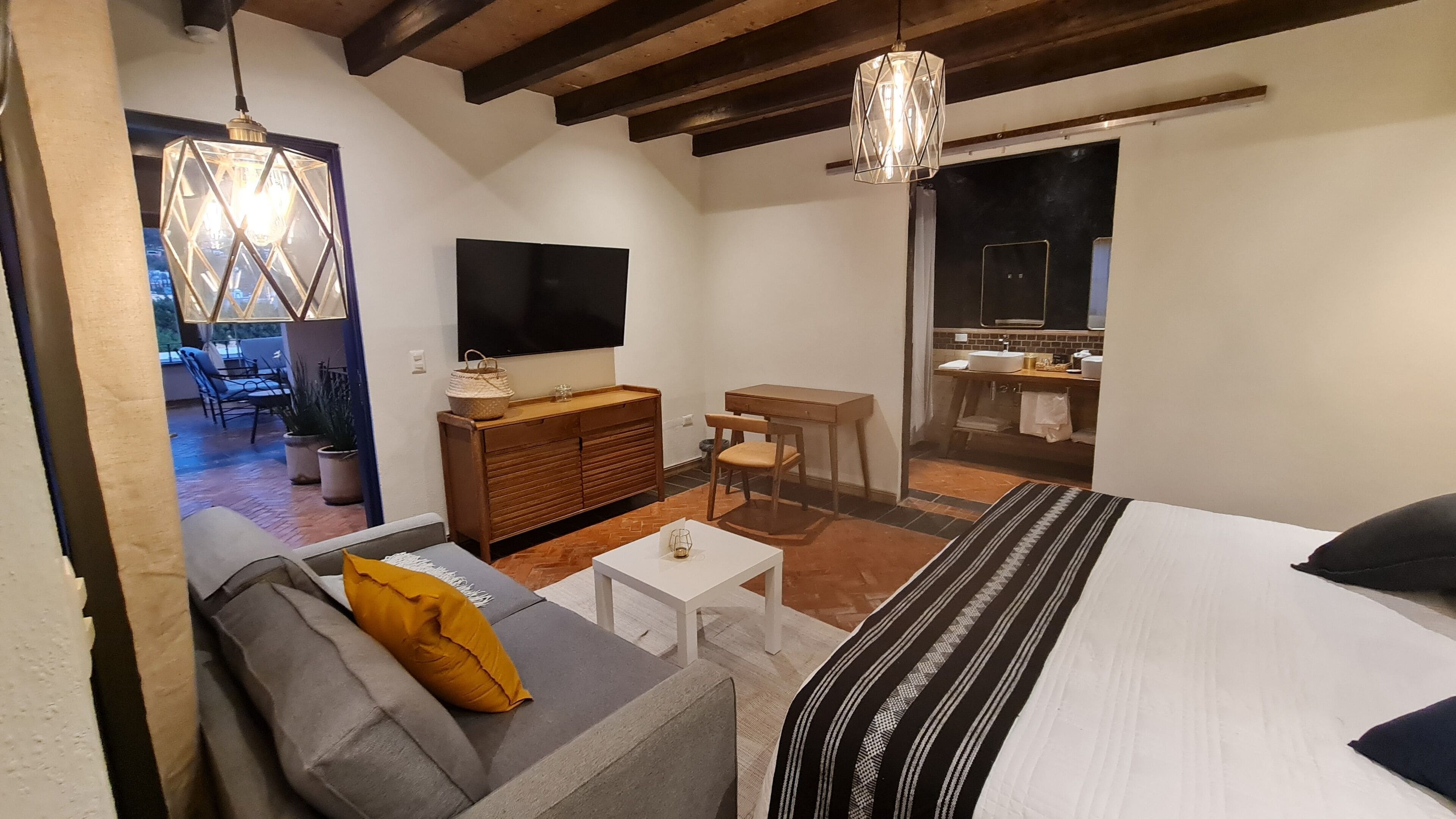 A cozy hotel room with a bed, a sofa with a yellow pillow, a TV, a wooden dresser, a small table, and a bathroom visible through an open door.