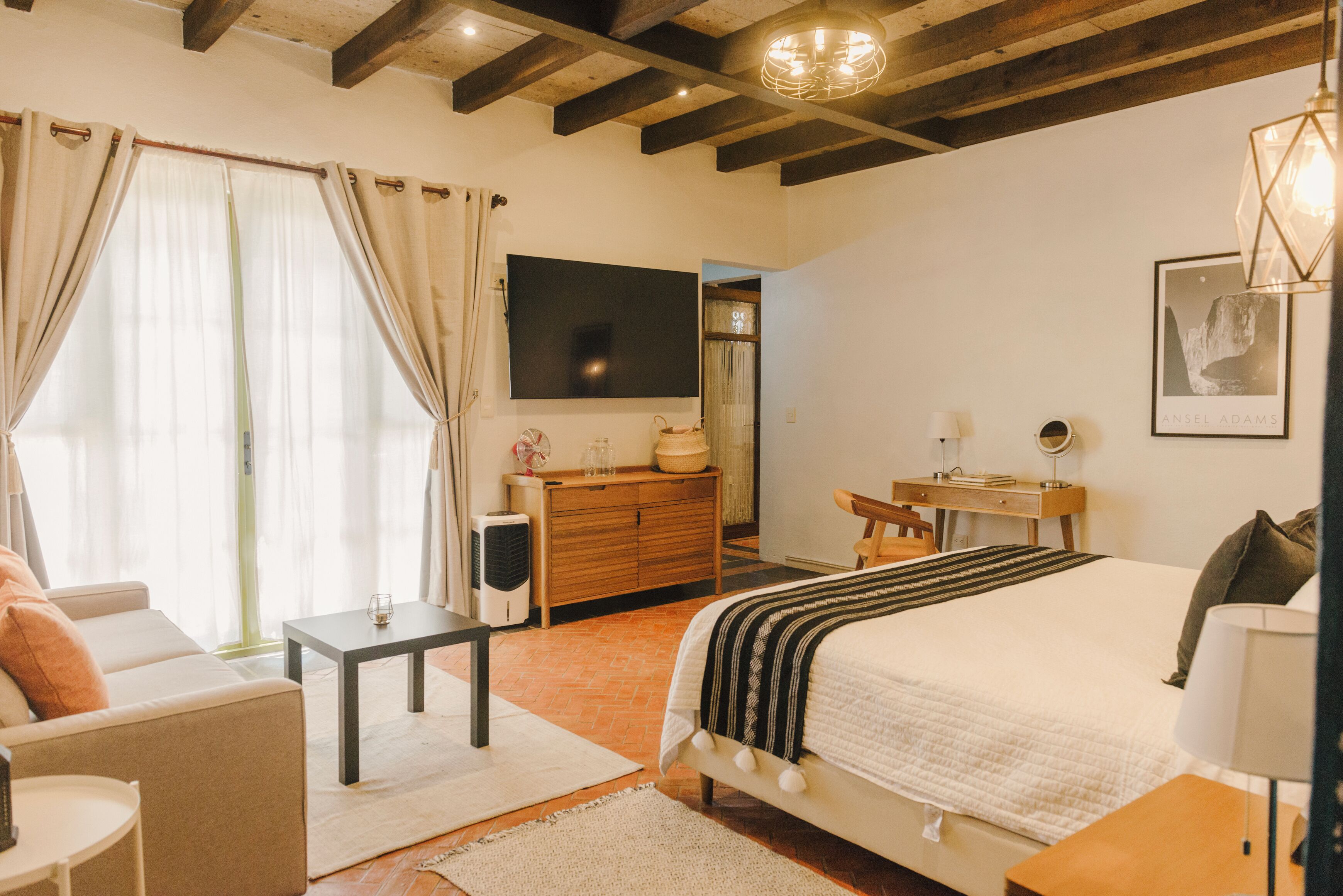 Une chambre bien éclairée, avec un lit, une table de chevet et un bureau avec une chaise. Un téléviseur est fixé au mur et un tableau encadré est accroché au-dessus du lit.