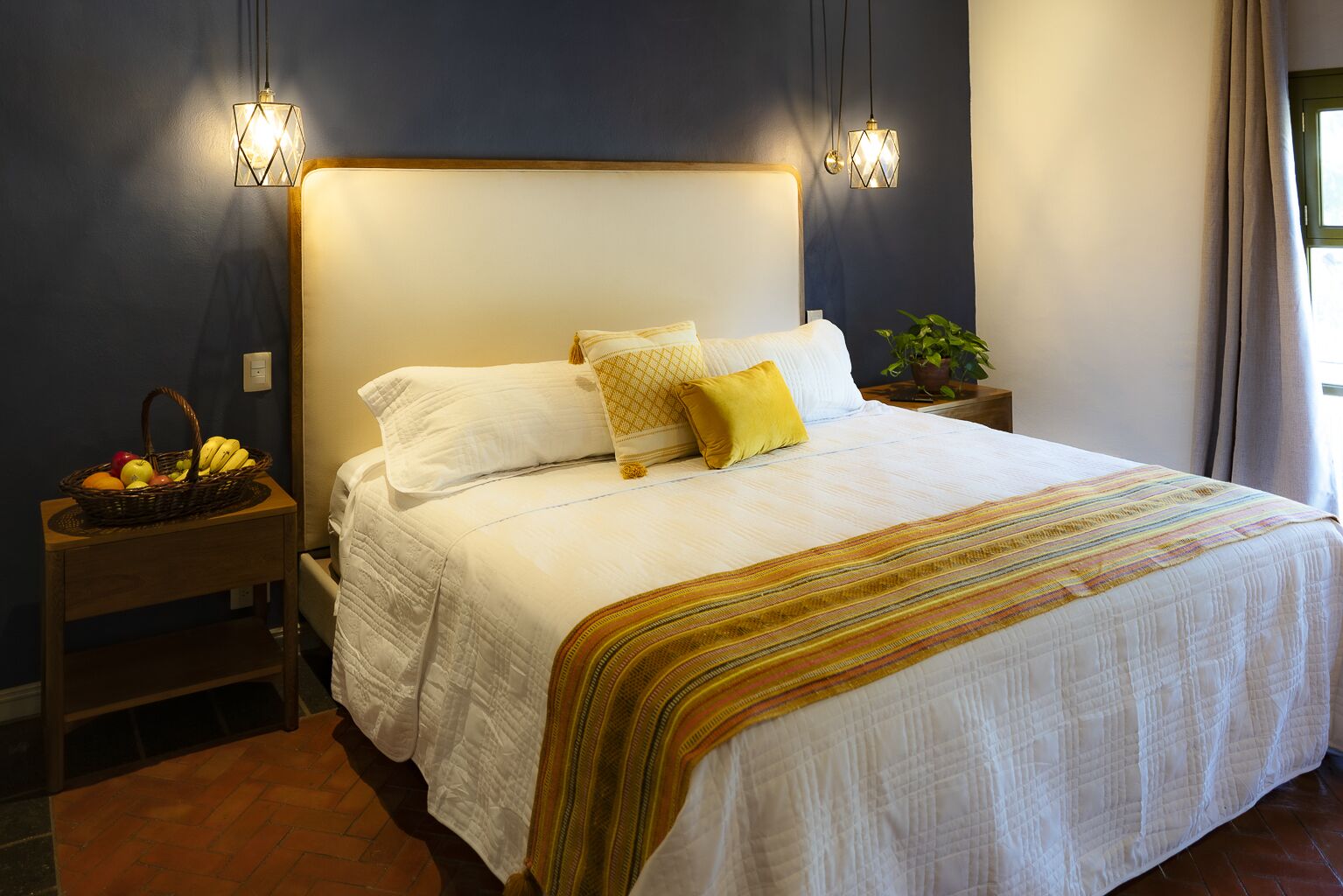A neatly made bed with white linens and a striped throw blanket, a bedside table with a fruit basket, and two wall-mounted lamps.