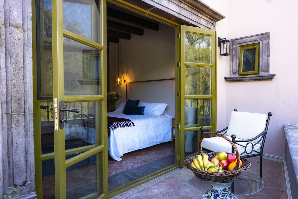 Une porte ouverte donne sur une chambre à coucher où se trouvent un lit, une chaise et un panier de fruits posé sur une table.