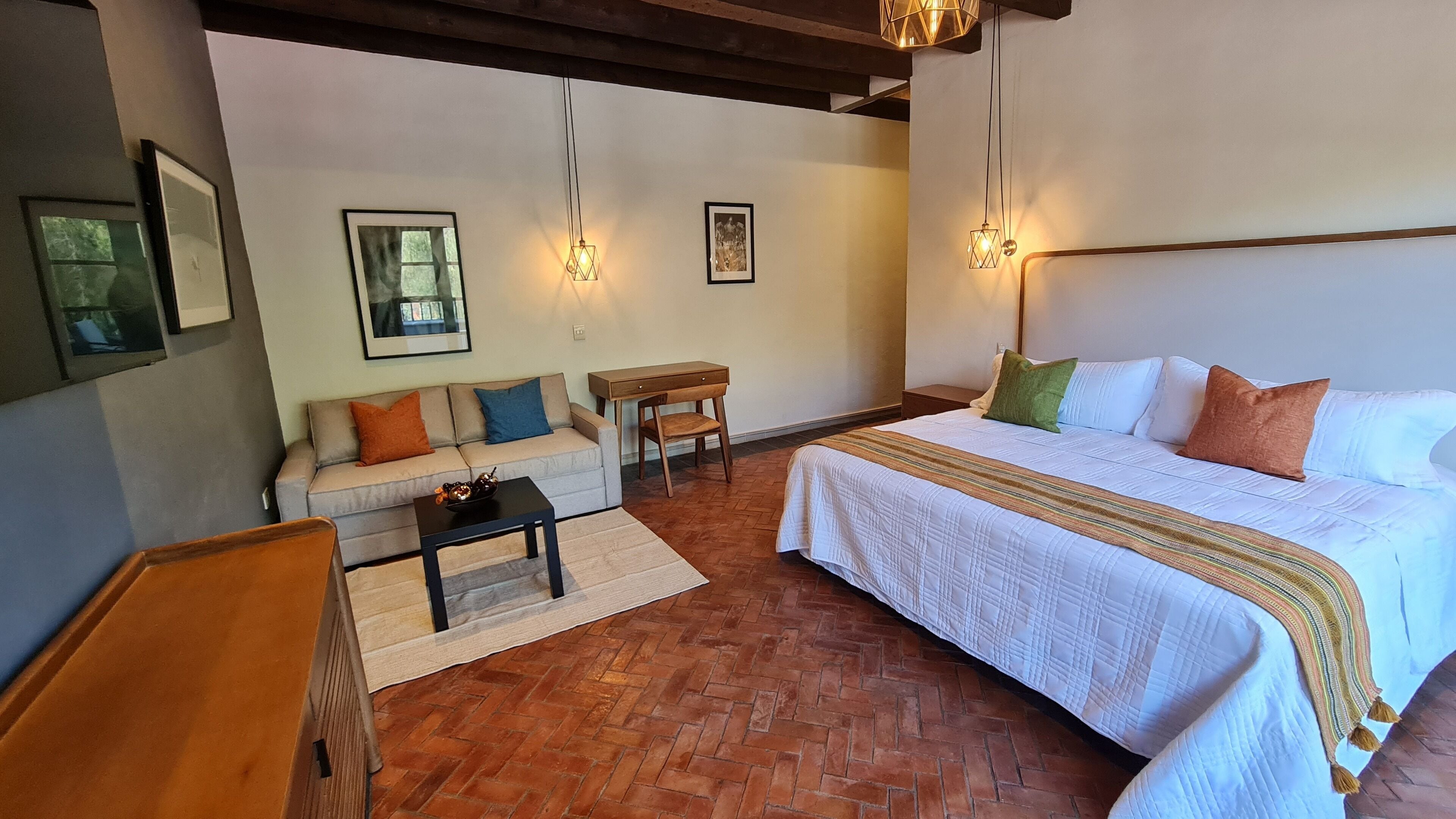 A cozy room with a bed, a sofa, a small table, and a chair. There are two wall-mounted lights and a picture on the wall.