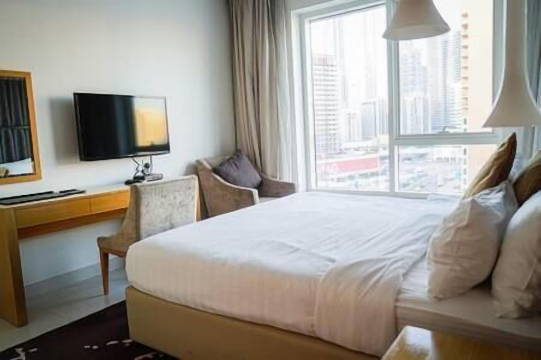 A hotel room with a large bed, a desk with a flat-screen TV, a chair, and a view of the city through the window.