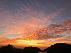A sunset over a hilly landscape with silhouettes of trees and a distant structure.