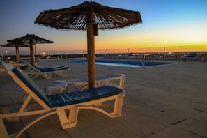 A rooftop pool area with lounge chairs and umbrellas, a swimming pool, and a view of the city skyline at dusk.