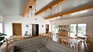A spacious room with a large woven sofa, wooden tables, and chairs. There are wooden beams on the ceiling and a view of the outdoors through large windows. - Monte Sardinha (Santiago do Cacém)