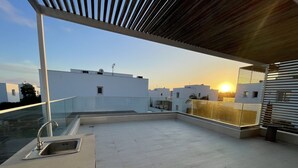Terraza en la azotea con un lavamanos moderno y barandilla de cristal con vistas a un paisaje urbano al atardecer.