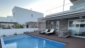 Una piscina con muelle de madera, sillas de descanso y una zona de estar cubierta.