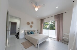 Relax in this serene bedroom with a large bed and ceiling fan, surrounded by a minimalist design and natural light.